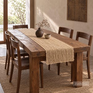 May include: A rectangular woven jute table runner with a textured, open-weave pattern and scalloped edges, centered on a rustic wooden dining table. The runner is light tan in color and features a consistent, natural fiber construction throughout.