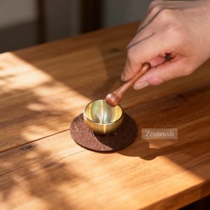May include: A small, gold-coloured singing bowl with a wooden mallet, resting on a brown felt pad. The bowl is held by a person's hand, suggesting its use in meditation or sound healing. The background is a wooden surface.