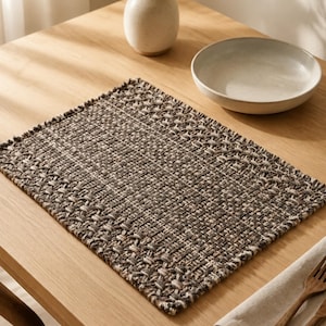 May include: A rectangular placemat featuring a thick, woven texture in a blend of dark brown, charcoal grey, and cream-coloured fibres. The edges of the placemat have a frayed, unfinished appearance, showcasing the intricate interlaced pattern.