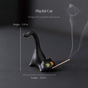 May include: A black cat-shaped incense holder with a green eye and an orange eye, holding a stick of incense. The text reads Playful Cat, Designed for standard stick incense, Height 3.35 in, Length 2.24 in. The cat has a long, curved tail.