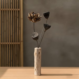 Op de afbeelding: Decoratief bloemstuk met gedroogde lotusbloemen en zaaddozen. De opstelling wordt gepresenteerd in een rustieke, cilindrische houten vaas. De lotusbloem heeft een metallic gouden kleur en de zaaddozen zijn donkerbruin.