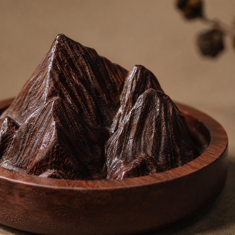 May include: A dark brown wooden sculpture shaped like a mountain range with jagged peaks and deep crevices, set inside a circular wooden bowl. The wood grain is visible throughout the textured surface, showing natural patterns and dark brown tones.