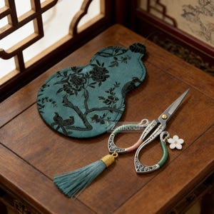 May include: Decorative embroidery scissors with a teal tassel and a white flower charm. The scissors have ornate silver handles and a teal and pink accent. A teal fabric case with a black floral design is also present.