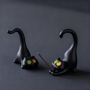 May include: Two black cat figurines with arched tails and wide, mismatched eyes in yellow and green. One cat is in a crouching position with a thin brown incense stick held between its front paws, while the other cat rests nearby.