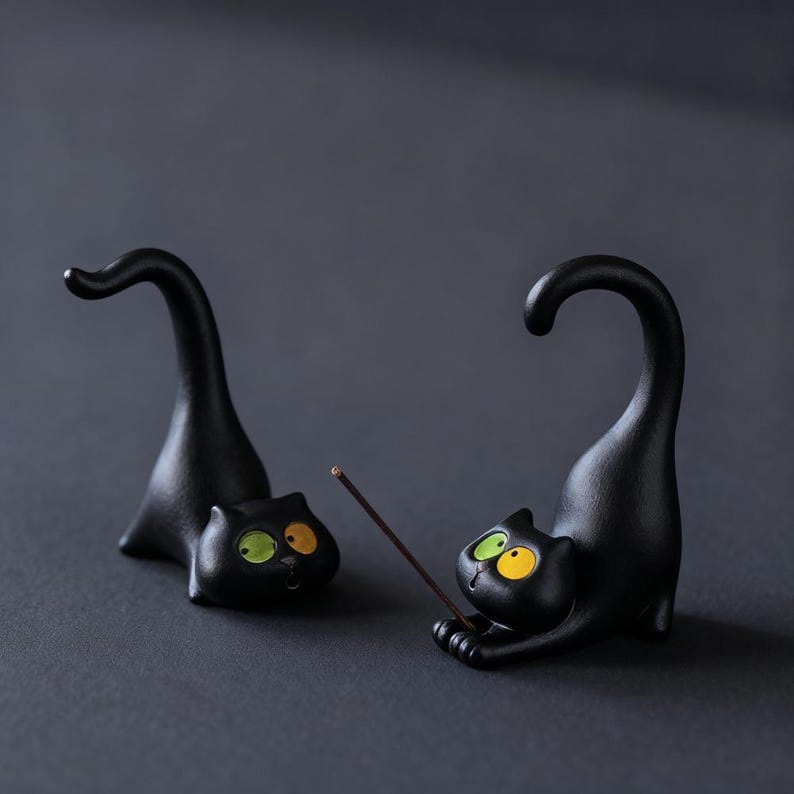May include: Two black cat figurines with arched tails and wide, mismatched eyes in yellow and green. One cat is in a crouching position with a thin brown incense stick held between its front paws, while the other cat rests nearby.