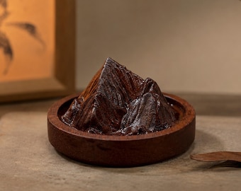 May include: A dark brown wooden sculpture shaped like a mountain range, set within a circular wooden base. The sculpture features carved ridges and peaks with a polished finish, creating a textured appearance that mimics a natural landscape formation.