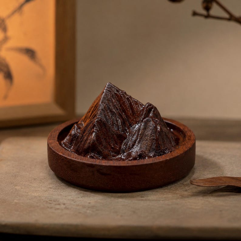 May include: A dark brown wooden sculpture shaped like a mountain range, set within a circular wooden base. The sculpture features carved ridges and peaks with a polished finish, creating a textured appearance that mimics a natural landscape formation.