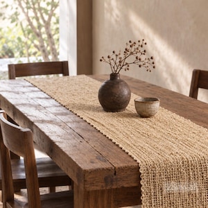 May include: A tan, open-weave table runner with a textured, grid-like pattern. The runner is laid flat across a dark brown wooden table, featuring a rounded dark brown vase containing dried branches and a small, speckled ceramic bowl.