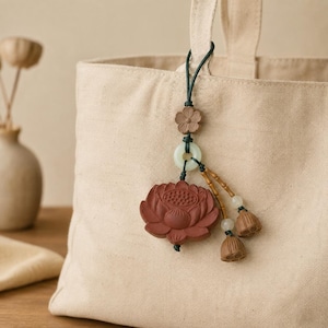 May include: A decorative bag charm featuring a terracotta-colored lotus flower pendant, a small carved flower, a pale green jade-like ring, and two dangling brown seed pod beads, all suspended from a dark green cord attached to a beige canvas tote bag.