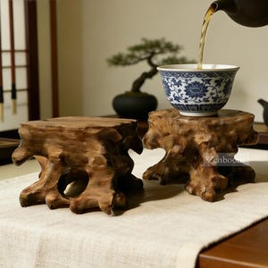 Peut inclure: Deux supports de présentation en racine de bois naturel avec des textures organiques noueuses et une surface supérieure carrée. L'un des supports soutient un bol à thé en porcelaine blanche orné de motifs floraux bleus, où du thé est versé. Texte : Zenboodi.