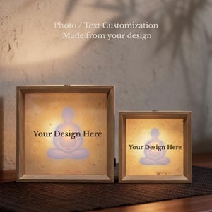 Puede incluir: Dos cajas de luz cuadradas de madera con iluminaci&oacute;n interna. Cada caja cuenta con un panel transl&uacute;cido que muestra la silueta de una persona en posici&oacute;n de meditaci&oacute;n sentada y el texto "Your Design Here" en letras negras.