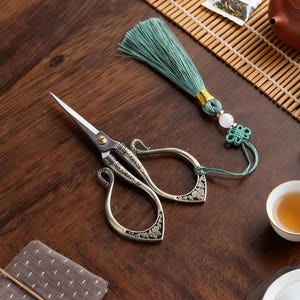 May include: Ornate silver scissors with a detailed floral design, paired with a teal tassel and decorative knot. The scissors are on a dark wooden surface, suggesting a tea ceremony or craft setting.