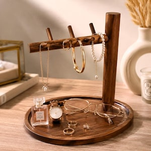 May include: A dark wood jewelry organizer stand with a circular base and a horizontal bar featuring three pegs. The stand holds a gold chain necklace, a gold bangle, and a white beaded bracelet. The base holds a perfume bottle, a watch, glasses, and rings.