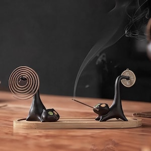 May include: Two black cat-shaped incense holders on a rectangular bamboo tray. One cat holds a brown spiral incense coil, while the other holds a thin incense stick with smoke rising. A small round metal coin hangs from the second cat's tail.