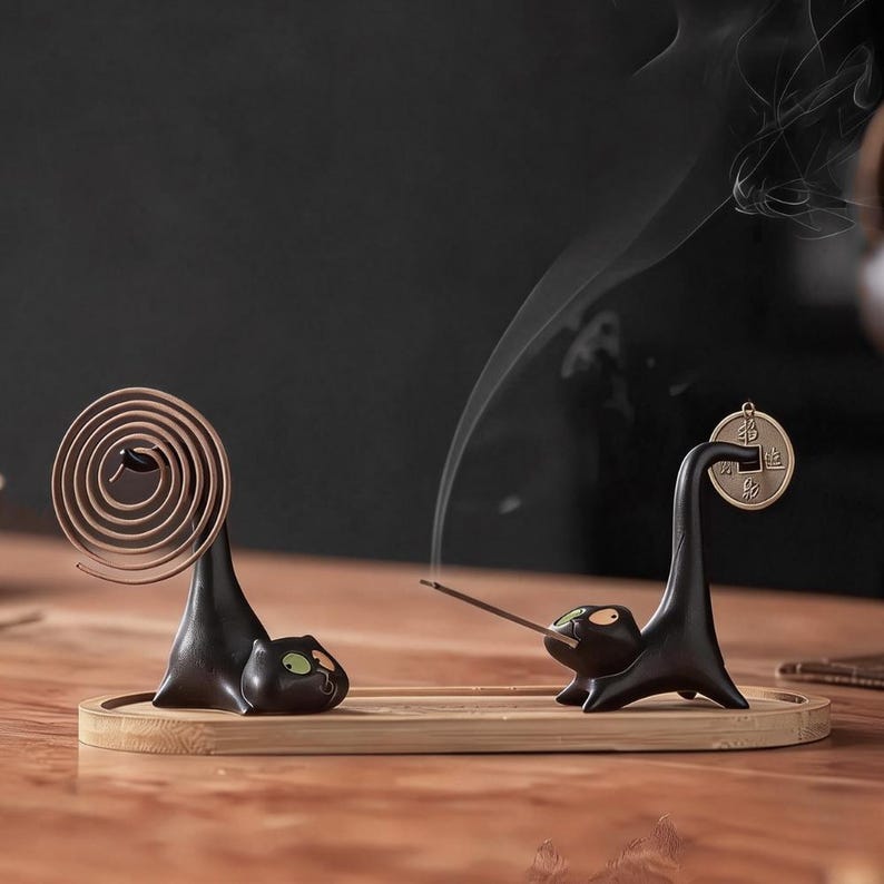 May include: Two black cat-shaped incense holders on a rectangular bamboo tray. One cat holds a brown spiral incense coil, while the other holds a thin incense stick with smoke rising. A small round metal coin hangs from the second cat's tail.