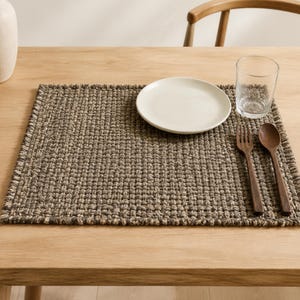 Puede incluir: Un mantel individual rectangular con una textura tejida gruesa en tonos marrón oscuro y gris. Sobre el mantel hay un plato de cerámica blanca, un vaso de cristal transparente, un tenedor de madera oscura y una cuchara de madera a juego.