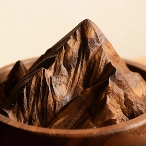 K&ouml;nnte beinhalten: Nahaufnahme einer Holzberg-Schnitzerei. Die dunkelbraune Schnitzerei zeigt eine Bergkette mit strukturierten Graten und Gipfeln, eingebettet in einer runden Holzschale. Die Holzmaserung ist sichtbar und tr&auml;gt zur nat&uuml;rlichen &Auml;sthetik bei.