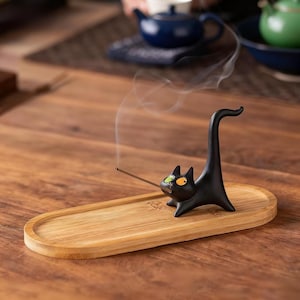 May include: A black ceramic cat incense holder with one green eye and one orange eye, featuring an upright tail. The cat is positioned on an oval bamboo tray, holding a thin, lit incense stick that emits a trail of white smoke into the air.