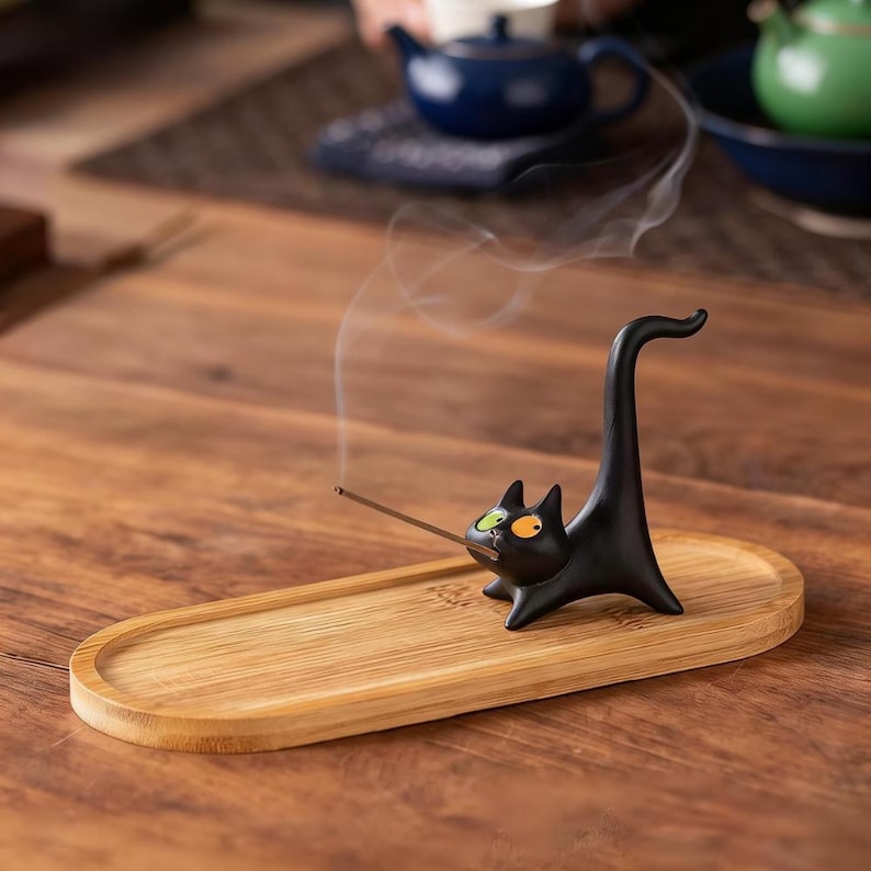 May include: A black ceramic cat incense holder with one green eye and one orange eye, featuring an upright tail. The cat is positioned on an oval bamboo tray, holding a thin, lit incense stick that emits a trail of white smoke into the air.