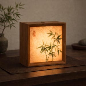 Op de afbeelding: Een vierkante houten lamp met een bamboe-ontwerp op het voorpaneel. De lamp geeft een warme gloed, en de bamboe-illustratie is in groen- en bruintinten. De lamp staat op een donkerbruin oppervlak.