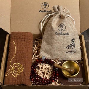 Op de afbeelding: Een geschenkdoos met een bruine handdoek met gouden borduursel, een kralenarmband, een kleine zangkom met een houten hamer en een zakje met trekkoord. De doos en de tas hebben de tekst "ZenBoodi".