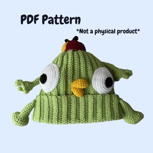 Puede incluir: Un gorro verde tejido a ganchillo con grandes ojos blancos, un pico amarillo y brazos pequeños. El gorro tiene el texto "PDF Pattern" y "*Not a physical product*".