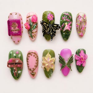 May include: A set of ten press-on nails featuring 3D designs including a pink book titled The Book of Fairies, tulips, a black butterfly, a fairy, pink blossoms, a brown sign reading Fairy Garden, a gold necklace, a yellow flower, and wings.