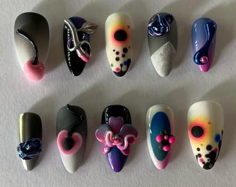 以下が含まれることがあります： ユニークなデザインの人工爪10個のコレクション。各爪は、さくらんぼ、抽象的な形、花の装飾など、異なるデザインが特徴です。色は黒、白、ピンク、青、紫で、さまざまなテクスチャと仕上げが施されています。