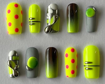 Op de afbeelding: Een set van tien kunstnagels in verschillende ontwerpen. De nagels hebben neongele, grijze en bruine kleuren. Sommige nagels hebben roze stippen, zilveren accenten of een gradiënteffect. De nagels zijn vierkant van vorm.