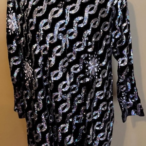 May include: A long black coat with a velvet texture, adorned with a repeating silver sequin pattern. The coat features long sleeves and a button-down front, with floral sequin embellishments.
