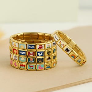 May include: Gold-toned charm bracelets with colorful enamel designs. One bracelet is upright, while the others are stacked. The charms feature various images and words, including "DREAM," "MOM," and a rainbow.