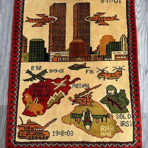 Op de afbeelding: Een beige tapijt met een rode rand en franjes. Het tapijt toont een stadsgezicht met twee torens en vliegtuigen, samen met kaarten en militaire voertuigen. Tekst bevat "9-11-01", "Iraq War" en andere data en locaties.