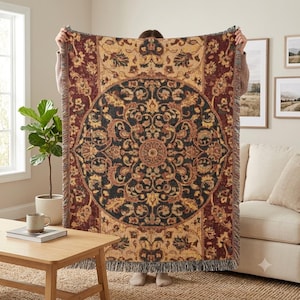 May include: A woven throw blanket featuring a large central circular medallion with intricate floral and scrollwork patterns in shades of deep red, black, gold, and cream. The rectangular blanket has a fringed edge on the top and bottom sides.