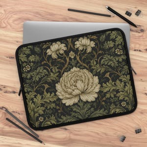 Op de afbeelding: Een zwarte laptoptas met een grote, crèmekleurige pioenroos en kleinere bloemaccenten. Het ontwerp bevat groene bladeren en wijnranken tegen een donkere achtergrond. De tas heeft een zwarte rits en zwarte rand.