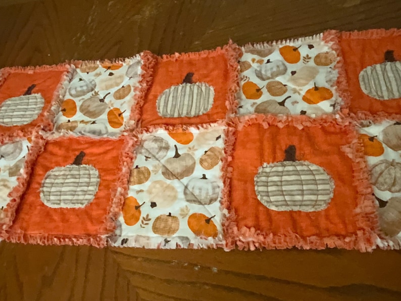 Pode incluir: Um caminho de mesa tipo rag quilt com um design de patchwork de quadrados laranja s&oacute;lidos alternados com quadrados brancos estampados com v&aacute;rias ab&oacute;boras. As bordas de cada quadrado s&atilde;o desfiadas, criando uma apar&ecirc;ncia texturizada.