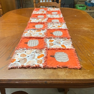 Pode incluir: Um caminho de mesa tipo rag quilt com um design de patchwork de quadrados laranja s&oacute;lidos alternados com quadrados brancos estampados com ilustra&ccedil;&otilde;es de ab&oacute;boras. Os quadrados de tecido t&ecirc;m costuras desfiadas e expostas nas bordas.