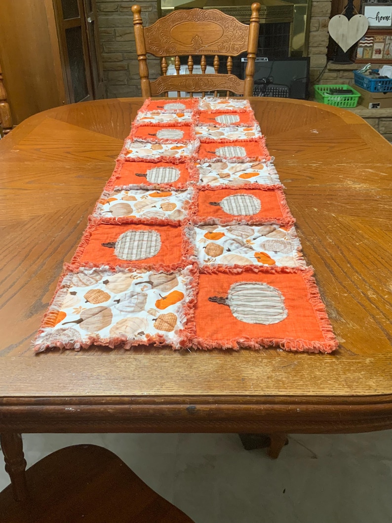 Pode incluir: Um caminho de mesa tipo rag quilt com um design de patchwork de quadrados laranja s&oacute;lidos alternados com quadrados brancos estampados com ilustra&ccedil;&otilde;es de ab&oacute;boras. Os quadrados de tecido t&ecirc;m costuras desfiadas e expostas nas bordas.