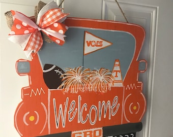 Puede incluir: Colgador de puerta de madera pintado de naranja y blanco en forma de camión con las palabras "Welcome" y "GBO" en él. El camión tiene un balón de fútbol, pompones y una bandera que dice "VOLS" en ella.