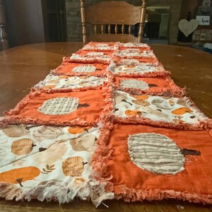 Pode incluir: Um caminho de mesa retangular estilo rag quilt com quadrados alternados de tecido laranja s&oacute;lido com aplica&ccedil;&otilde;es de ab&oacute;boras e tecido branco estampado com um padr&atilde;o de ab&oacute;boras e folhas em tons de laranja e bege, com bordas desfiadas.