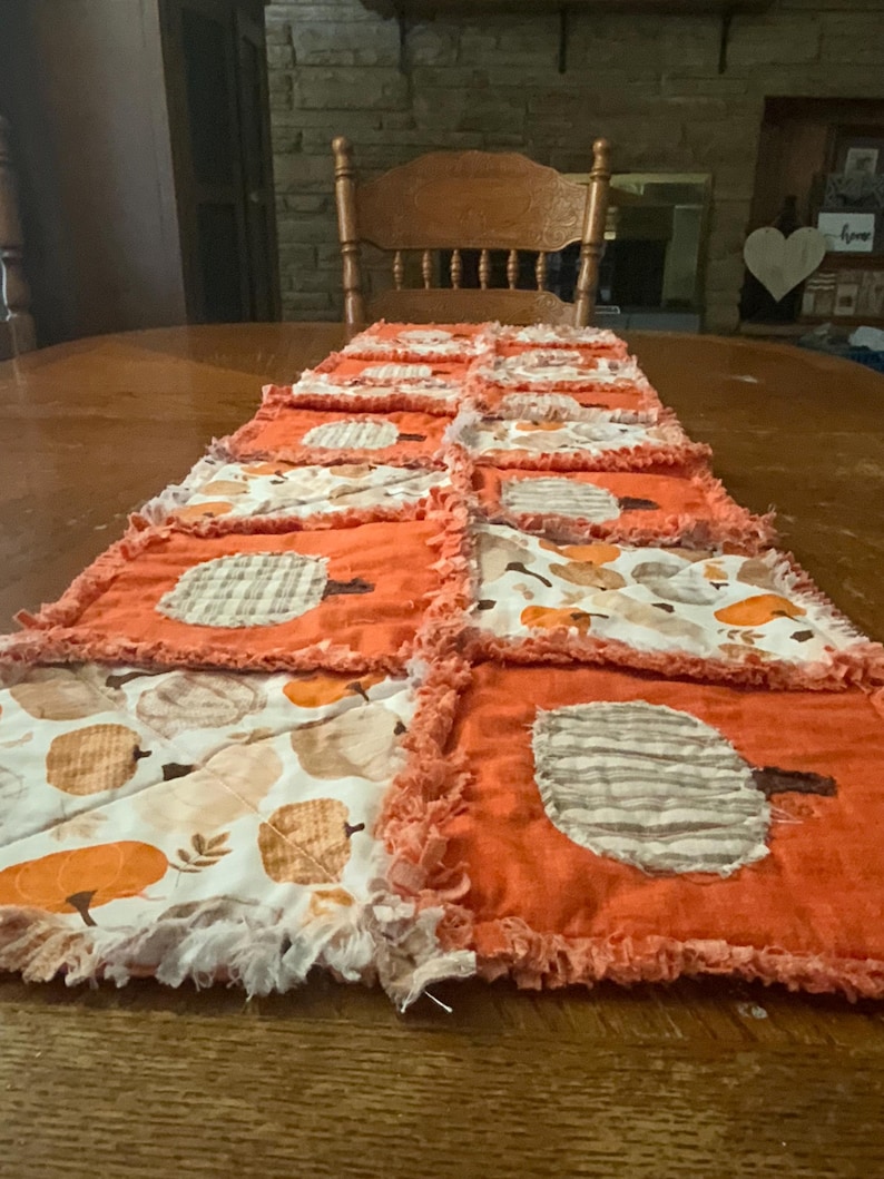 Pode incluir: Um caminho de mesa retangular estilo rag quilt com quadrados alternados de tecido laranja s&oacute;lido com aplica&ccedil;&otilde;es de ab&oacute;boras e tecido branco estampado com um padr&atilde;o de ab&oacute;boras e folhas em tons de laranja e bege, com bordas desfiadas.
