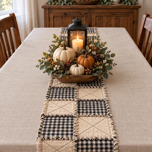May include: A rectangular table runner featuring a patchwork design of black and white buffalo check squares alternating with solid beige quilted squares, finished with frayed edges. A wooden bowl holds pumpkins and a black lantern.