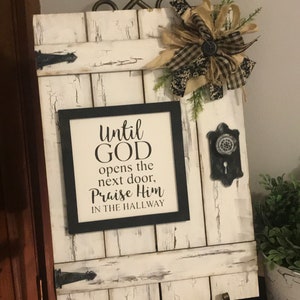 May include: A distressed white wooden wall decor piece with a black framed sign that reads "Until God opens the next door, Praise Him in the hallway." It features a decorative bow, metal hardware, and hooks.