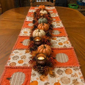 May include: A rectangular patchwork table runner featuring alternating squares of solid orange fabric and white fabric with a pumpkin print. The runner is decorated with a central arrangement of small artificial pumpkins, pinecones, and metal candle holders.