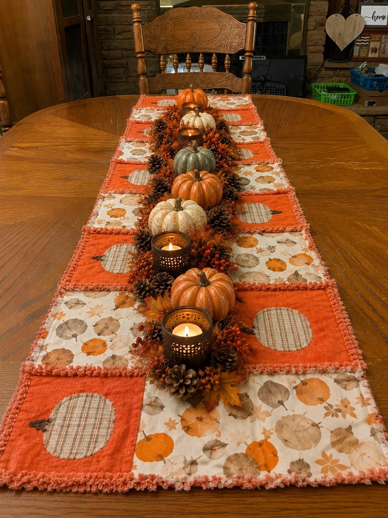 Pode incluir: Um caminho de mesa retangular de patchwork com quadrados alternados de tecido laranja s&oacute;lido e tecido branco com estampa de ab&oacute;boras. O caminho &eacute; decorado com um arranjo central de pequenas ab&oacute;boras artificiais, pinhas e casti&ccedil;ais de metal.