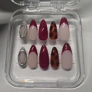May include: A set of ten artificial nails in a clear plastic case. The nails feature various designs, including a silver oval, a white tip with a burgundy base, solid burgundy, and a leopard print pattern. The nails are almond-shaped.