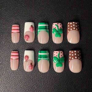 May include: A collection of ten false nails with diverse patterns. The nails showcase a nude base with designs such as pink and brown stripes, ladybirds, green plaid, green shamrocks, and brown polka dots. The nails are displayed on a dark surface.