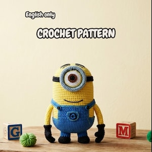 May include: A crocheted Minion toy in yellow and blue, with black accents. The toy has one large eye and is wearing blue overalls. The words "CROCHET PATTERN" are above the toy. A wooden block with the letter "G" is to the left.