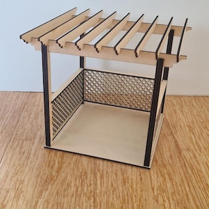 Puede incluir: Una pérgola de madera en miniatura con un lado de celosía y un techo de listones. La estructura está hecha de madera de color claro con ribetes marrón oscuro y está diseñada para fines decorativos. La pérgola se encuentra sobre una superficie de madera.