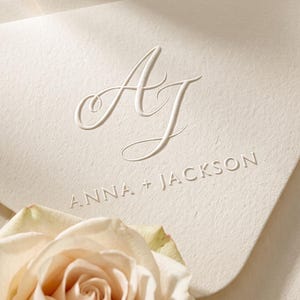 May include: A cream-colored envelope featuring an embossed monogram with the letters AJ in a cursive script, followed by the text ANNA + JACKSON in a clean, sans-serif font. A pale peach rose is positioned at the bottom of the envelope.
