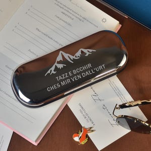 Könnte beinhalten: Ein silbernes Brillenetui mit einem Bergmotiv und dem Text "TAZZ E BCCHIR CHES MIR VEN DALL'URT". Das Etui liegt neben einem Notizbuch, einer Sonnenbrille und einem blauen Brillenetui.